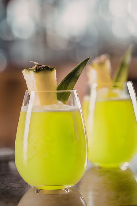Yellow-green tropical cocktail garnished with pineapple wedge and leaf, served in a clear glass.