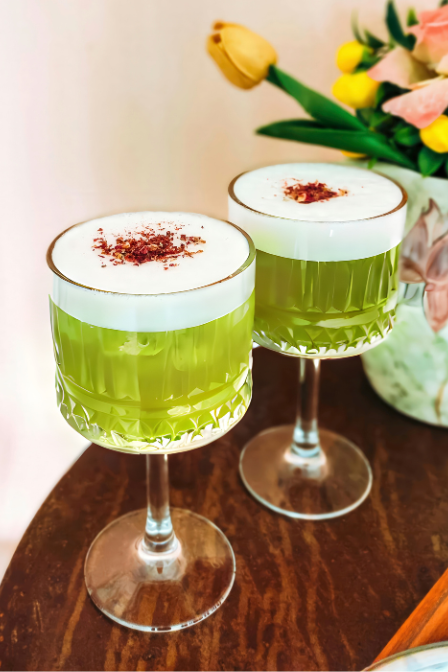 Two green drinks with foamy tops in elegant glasses on a table beside a vase of yellow flowers.